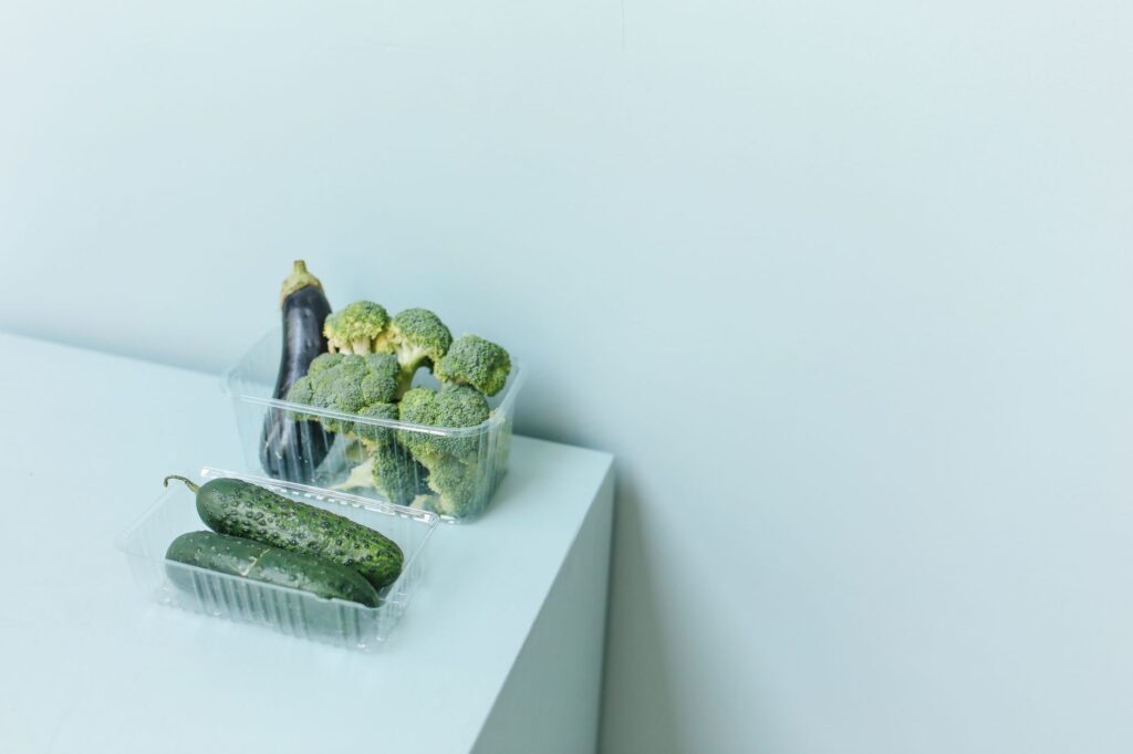 Assortment of fresh vegetables like broccoli, eggplant, and cucumber in plastic packaging on a white surface.
