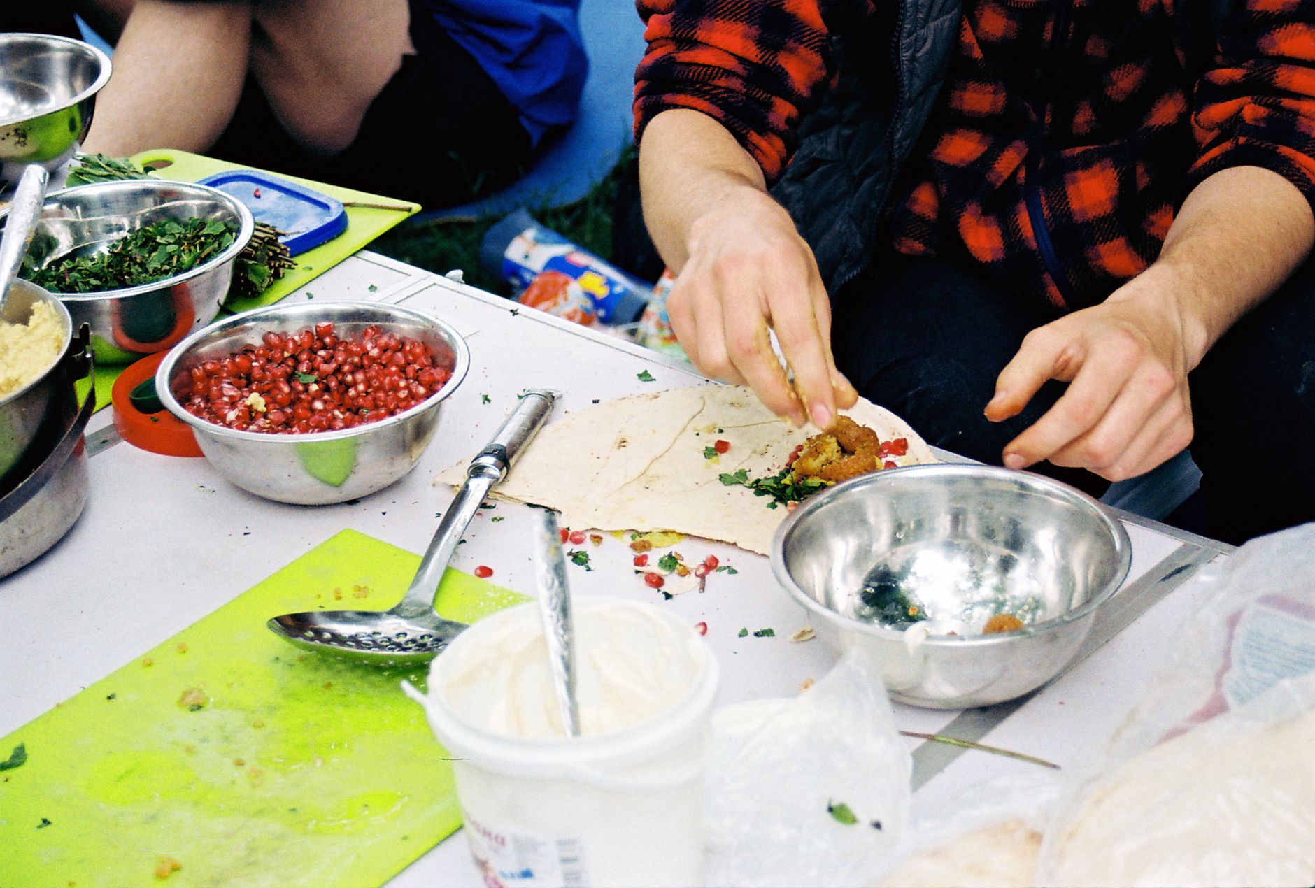 Hands preparing a falafel wrap with fresh ingredients like pomegranate seeds and herbs on a table.