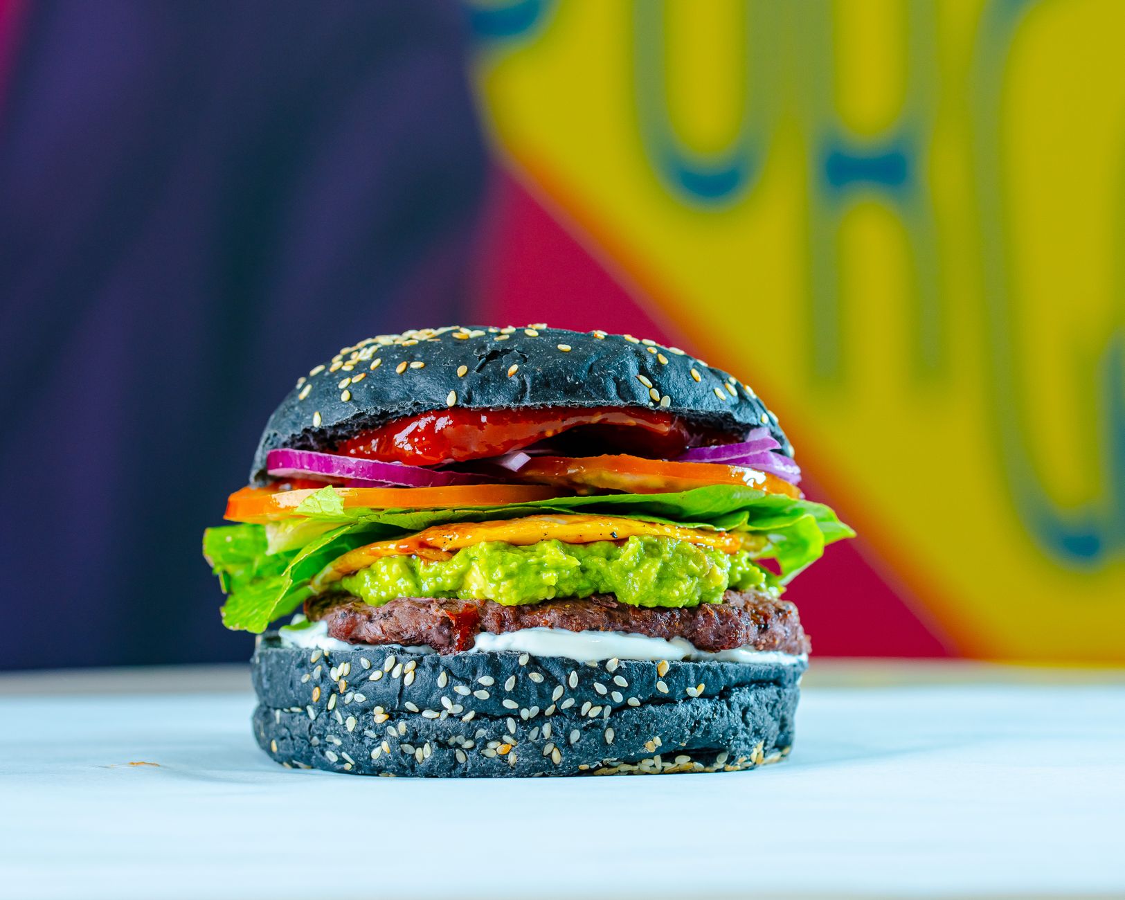 Colorful veggie burger with black sesame bun, avocado, and fresh veggies on a vivid background.