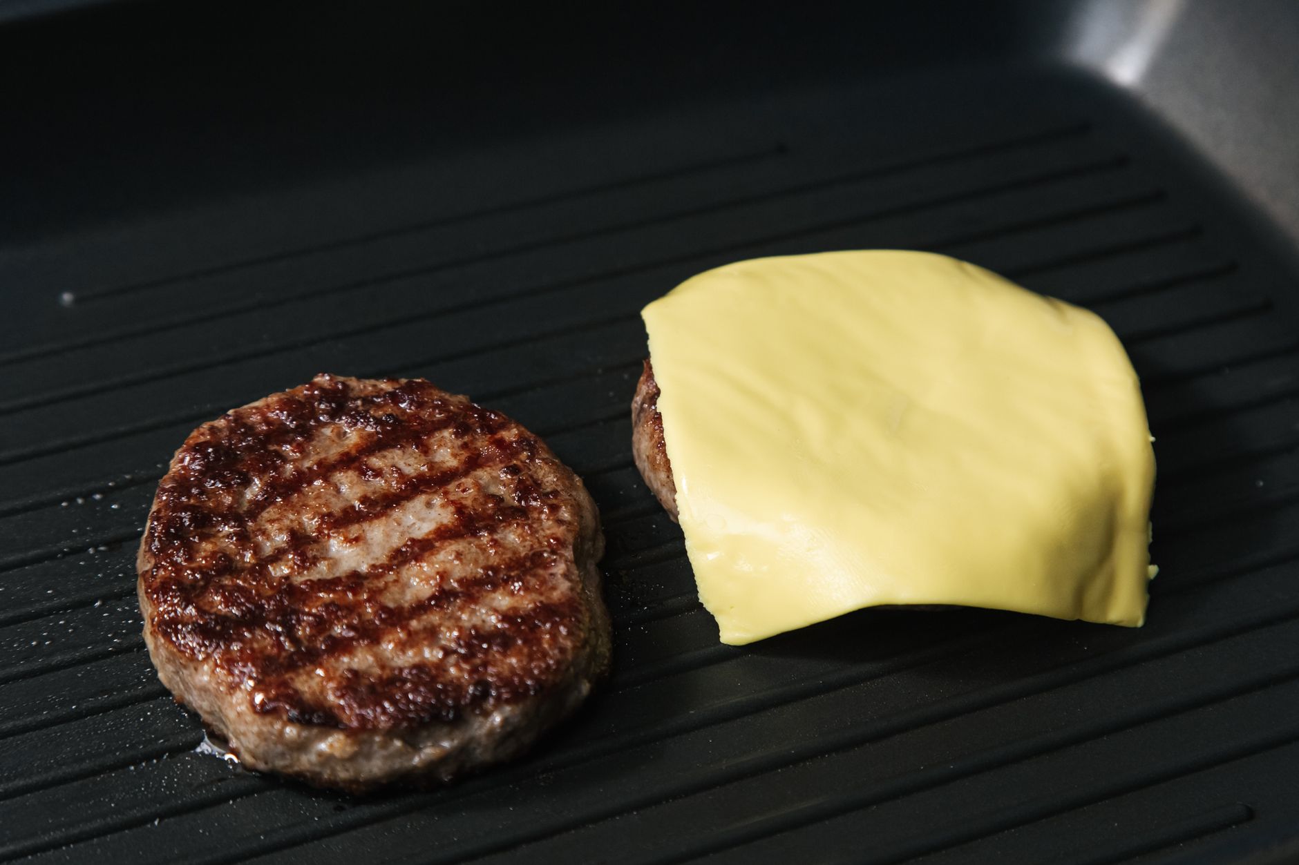 Close-up of grilled burger patties on a pan, one topped with melted cheese.