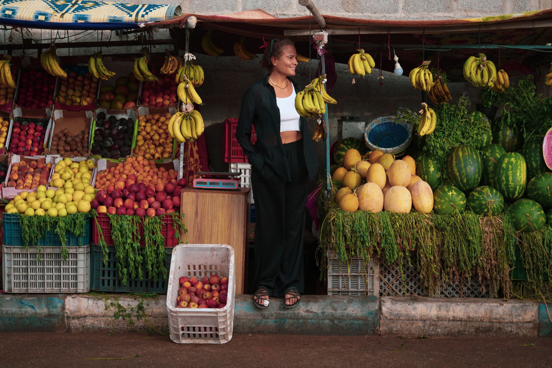 A woman stands by a colorful fruit stall in Oued Ed-Dahab with bananas, melons, and more.